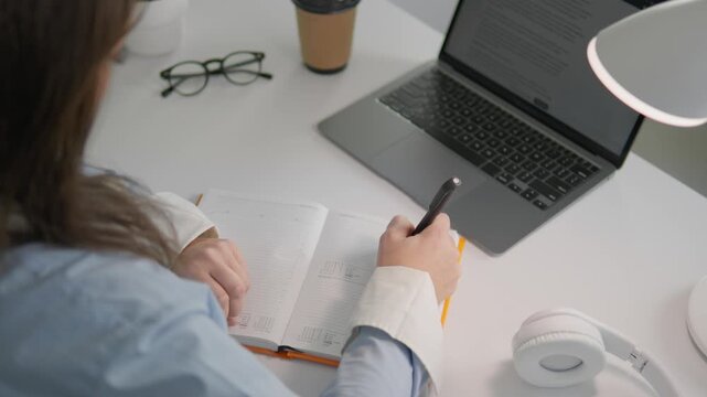 Focused female professional takes handwritten notes while using a laptop for remote work or online learning. Cozy workspace setup includes coffee, headphones, and organized stationery essentials