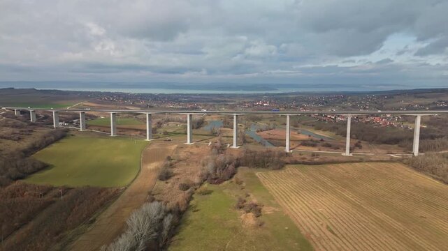 In Kőr&ouml;shegy, Hungary, the Kőr&ouml;shegyi V&ouml;lgyh&iacute;d spans a rural valley with open fields and nearby Lake Balaton, viewed from a drone perspective as vehicles move along the elevated highway.