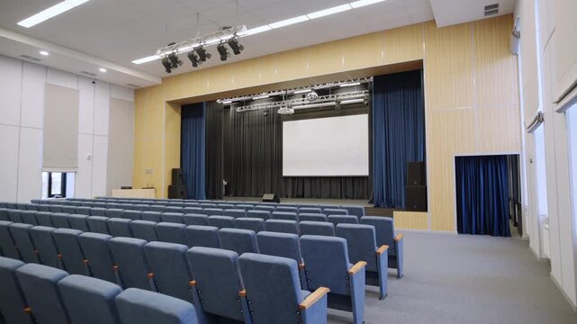 School assembly hall features rows of velvet seats facing large screen. Stage equipped with spotlights dark curtains and audio speakers