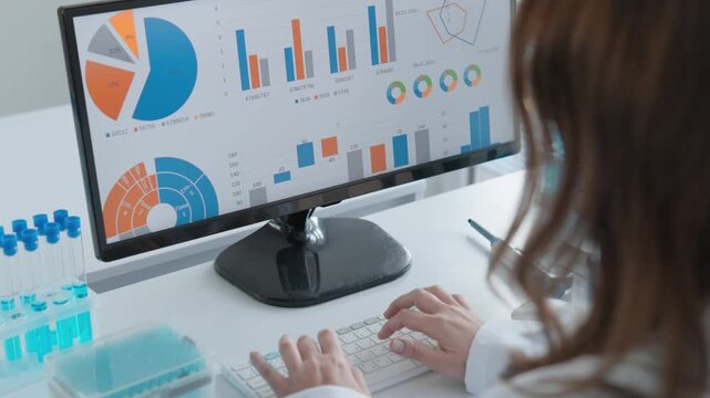 Woman in lab coat works at desk, reviewing scientific analytics, laboratory results on large monitor. Data visualization tools support innovation, research efficiency in laboratory environment