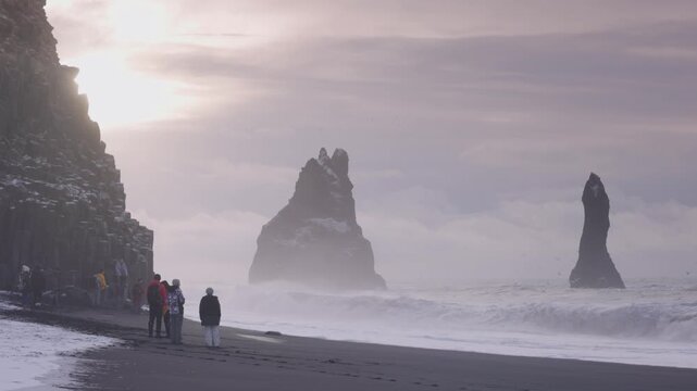 View of Reynisdrangar sea stacks on black sand beach with tourists standing on the shore and crashing waves in Iceland.
