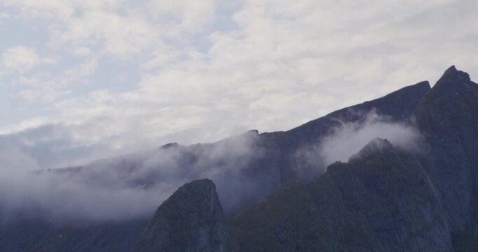 Craggy, jagged mountains rise under a cloudy sky, with mist swirling around their bases, creating a serene, ethereal, and mysterious atmosphere in a wild, untamed landscape.