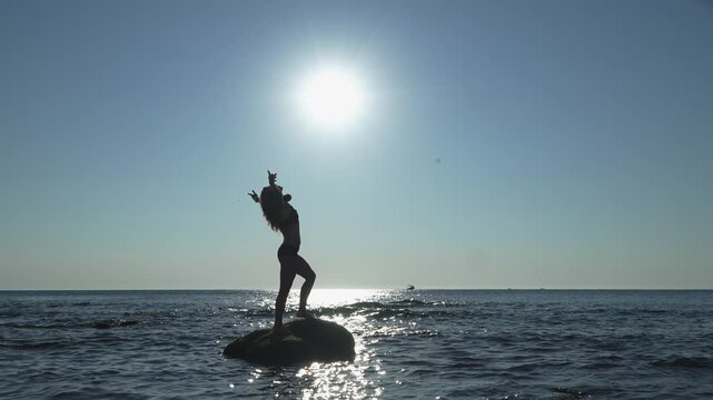 A carefree woman enjoys the ocean, standing on a rock with arms lifted, silhouetted by the glowing sun. The tranquil seascape and serene sky evoke feelings of freedom and happiness