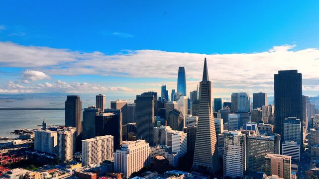 San Francisco financial district skyline with pyramid spire and glass tower. Aerial view over mid-rise rooftops, bay waterfront and Bay Bridge under blue daytime sky.
