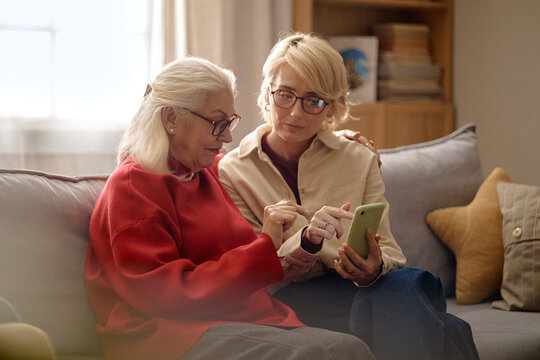 Medium shot of senior Caucasian woman using phone with middle aged Caucasian daughter, learning mobile app at home and supporting digital inclusion. Useful for tech training, family care marketing