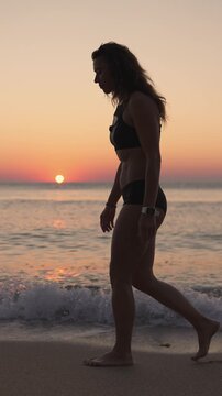 A young woman in sportswear strolls by the water's edge as the sun rises, casting a warm glow over the calm waves. The serene beach scene captures wellness, mindfulness, and summer vibes