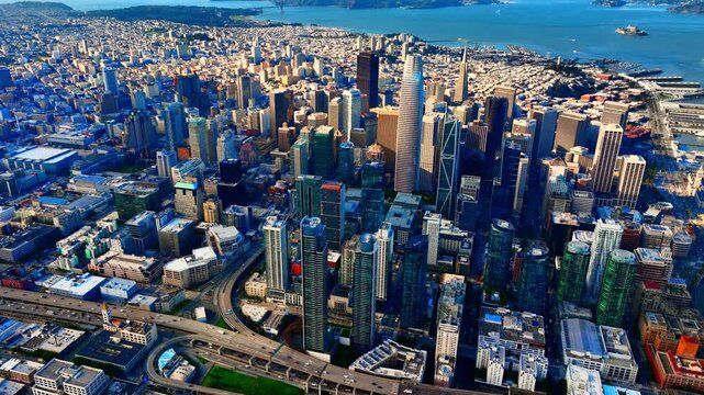 Panorama of San Francisco skyline with highway and bay beyond. Aerial view of dense city grid, tower cluster, curving freeway ramp and distant Golden Gate strait under blue sky.