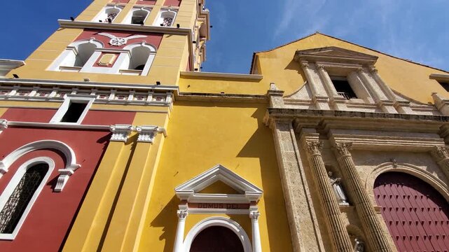 View of the colorful colonial church facade in the historic Getsemani neighborhood with vibrant yellow and red walls under a clear blue sky in Cartagena, Bolivar, Colombia.