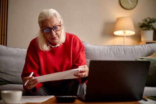 Medium shot of senior Caucasian woman reviewing paperwork and taking notes beside laptop at home, managing finances and online tasks. Useful for retirement planning, budgeting, remote admin