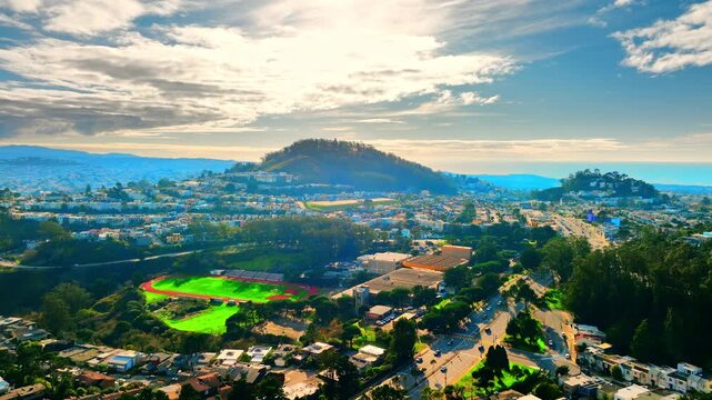 Elevated view of San Francisco hillside neighborhood with stadium track and Twin Peaks beyond. Green athletic field, red running track, tree lined streets and layered hill ridges under cloud sky.