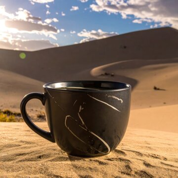 A steaming cup of coffee sits on a sand dune under a cloudy sky.