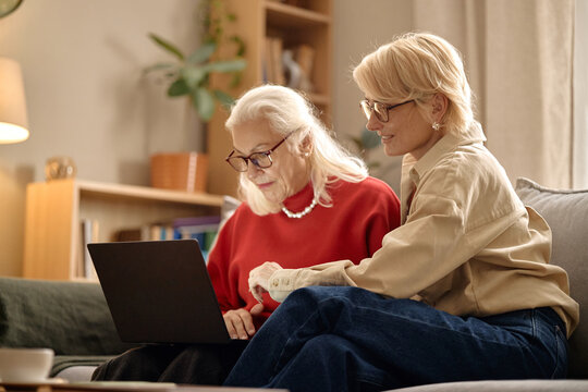 Medium shot of senior Caucasian woman using laptop while middle aged Caucasian woman assisting at home, supporting digital learning and family connection. Ideal for tech support or online service ads
