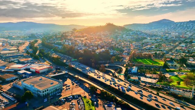 Sunset light over San Francisco freeway and hillside community toward East Bay mountains. Multilane highway, warm orange glow on rooftop grid, green park strip and hazy East Bay ridge at golden hour.