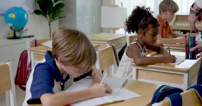 Elementary boy passes pencil case to male teacher then writes worksheet at wooden desk, multicultural classroom activity builds responsibility, attention, school routine, class participation