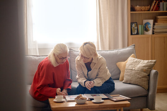Senior Caucasian woman and middle aged Caucasian woman reviewing photo album together at home, discussing family memories. Useful for caregiving, retirement marketing