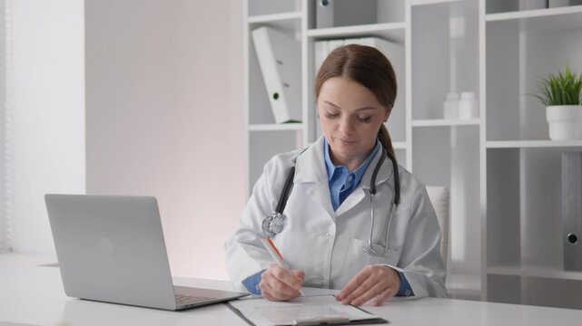 Young woman physician with stethoscope sits at bright workspace, filling out paperwork and updating patient files. Medical professional demonstrates dedication to healthcare and digital workflow