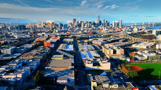 City street toward San Francisco downtown skyline and Bay Bridge at midday. Converging avenue, mixed use warehouse block, baseball field, high rise cluster and bay under clear blue sky.