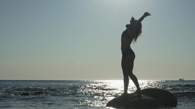 A young woman enjoys a peaceful moment on a rocky shore, silhouetted against the setting sun and sparkling sea. The scene radiates freedom, relaxation, and the beauty of nature in summer