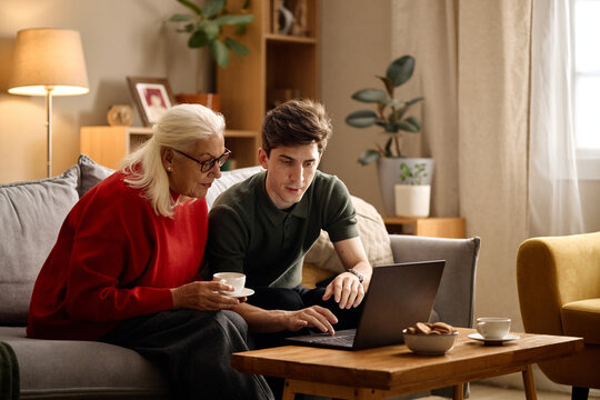 Senior Caucasian woman and young adult man reviewing laptop together at home, discussing online task and digital support. Useful for family tech help, banking, insurance, services