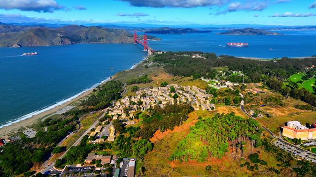 Coastal headland with Golden Gate Bridge view, San Francisco. Rugged cliffside community, sandy beach, red bridge span across blue bay and Marin hills under cloud-dotted sky.