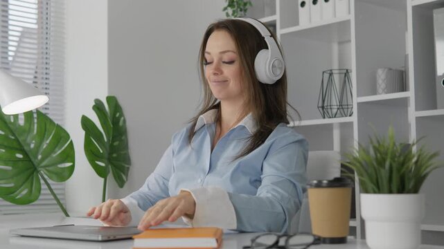 Professional woman in headphones listens to music at her bright office workspace, surrounded by plants and technology. She combines focus and relaxation, creating a positive work atmosphere