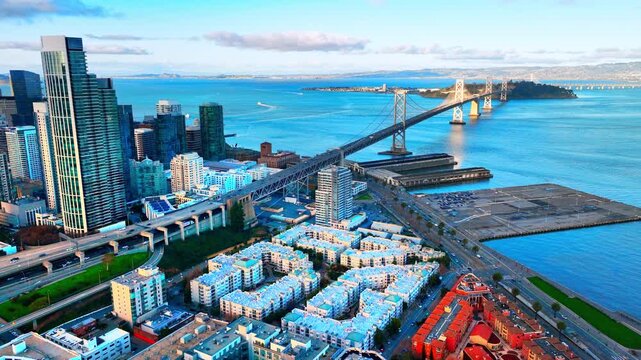 Downtown skyline and Bay Bridge from waterfront, San Francisco. Glass tower cluster beside bay, suspension bridge spanning to Yerba Buena Island and East Bay hills under partly cloudy sky.