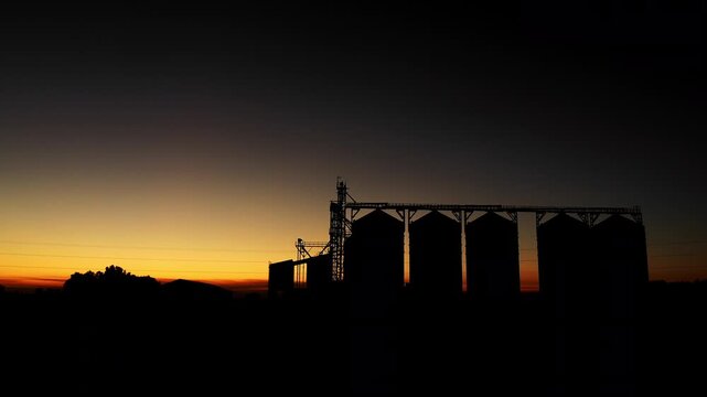 Wide view of grain elevator and storage silos silhouetted against colorful twilight sky. Agriculture, grain logistics and food supply chain concept.