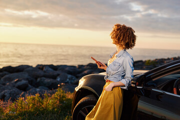 Fototapeta premium A woman with curly hair stands beside a car at the coast during sunset, looking at her phone. A candid lifestyle scene about travel, relaxation, and modern routines.