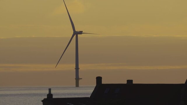 Telephoto dawn shot framing a massive, static wind turbine from the Blyth Offshore Demonstrator rising behind the silhouetted roofs of St Mary's Lighthouse cottages.
