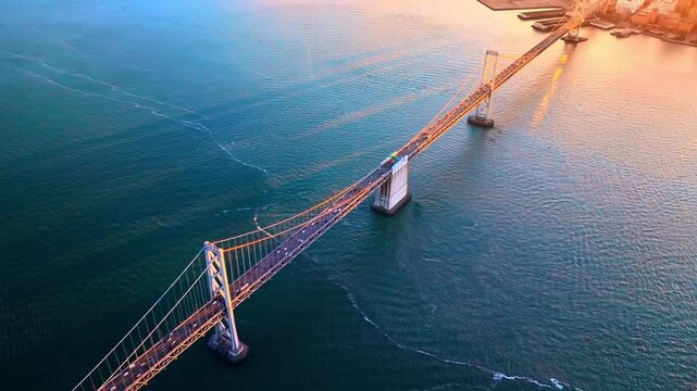Bay Bridge western suspension section at sunset, San Francisco Bay. Aerial view looks along twin tower span over teal water, golden warm light on cable array and city pier in upper corner.