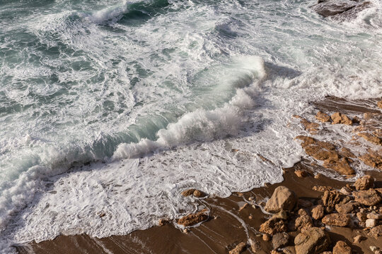 Rouleau d'une vague qui s&rsquo;&eacute;tale sur le sable et la roche brune, bord de mer agit&eacute;, gros plan, mousse blanche