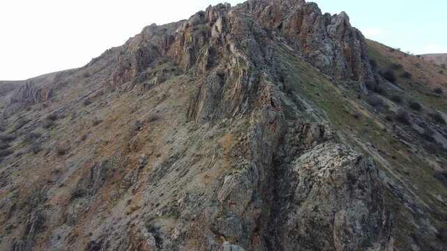 Drone shot flying towards a towering rocky summit with steep gravel slopes, stone outcroppings and dry grass patches in Kazakhstan. 