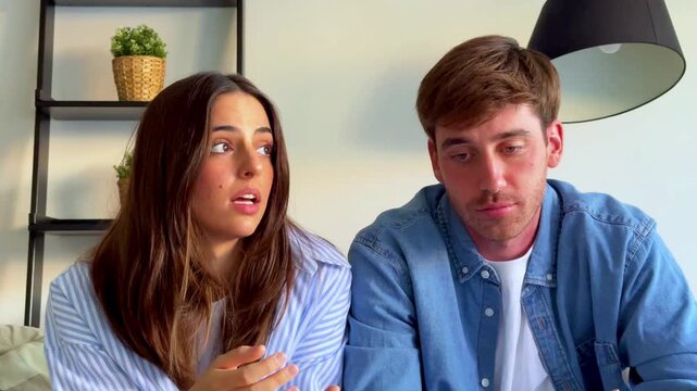 A young couple sitting together, engaged in an expressive conversation, showcasing a range of emotions and reactions. They are indoors in a modern home, perhaps discussing important matters or sharing