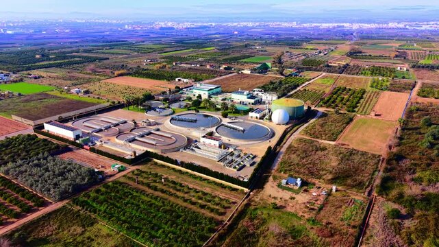 Aerial view of wastewater treatment plant amid farmland, Valencia, Spain. Drone perspective circular clarifier tank, white digester dome, green facility buildings and orange groves under clear sky.