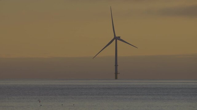 Super telephoto dawn shot of a rotating offshore wind turbine from the Blyth Offshore Demonstrator project positioned out at sea against a golden horizon.