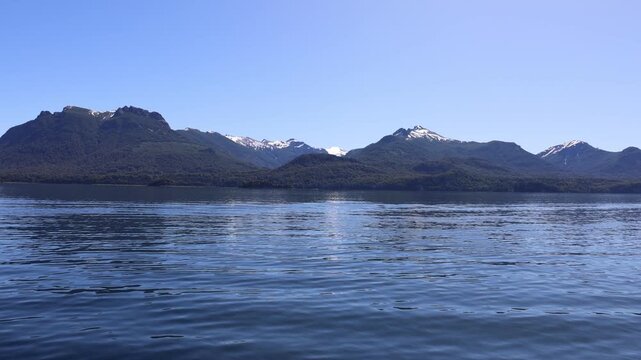 View of Isla Victoria with dense forests and snow-capped mountains across the calm blue waters of Nahuel Huapi Lake in San Carlos de Bariloche, Rio Negro Province, Argentina.