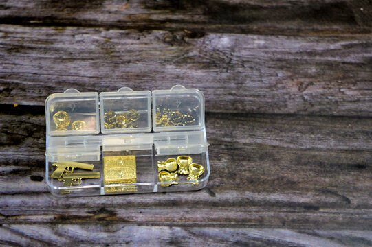 Small, clear plastic organizer box used for storing craft supplies or jewelry components, contains various small metal accessories, including gold-toned charms shaped like miniature guns, small rings