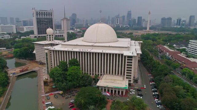 Wide aerial drone shot capturing Istiqlal Mosque in full with the Monas (National Monument) and Jakarta CBD skyline visible in the background.