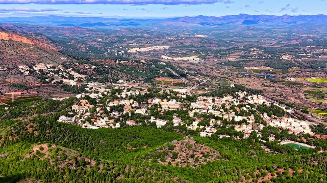 Aerial view of hilltop village amid forested valley and mountain range, Spain. White house cluster, pine canopy, winding road and distant rocky ridge under partly cloudy sky.