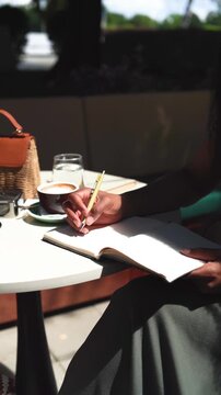 A black woman writes in a notebook paper diary closeup in a cafe on a sunny day outdoors