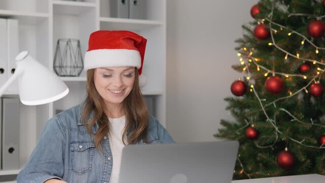 Cheerful female enjoying remote work in denim shirt and Santa hat, surrounded by holiday decorations. Christmas tree with red ornaments and warm lights creates joyful seasonal mood