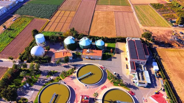 Aerial view of wastewater treatment facility with digester tank and solar panel, Spain. Circular clarifier, dome digester, silo tank, photovoltaic array and surrounding orange grove under daylight.