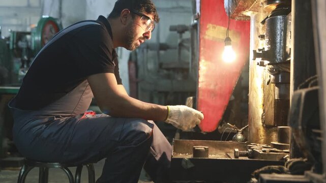 Male worker wearing safety glasses sits at his desk shaping circular aluminum sheets with a metal stamping machine illuminated only by dim light to ensure visibility during his work.