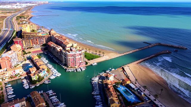 Coastal marina with colorful apartment buildings and sandy beach, Valencia, Spain. Teal harbor channel, moored boat, red tile rooftop, palm tree, breakwater, and turquoise sea under blue sky.
