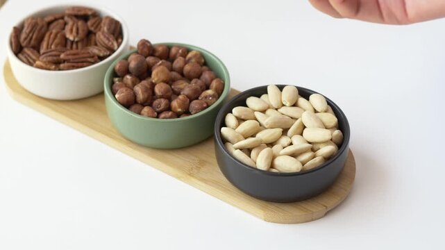 Fingers pick up almonds from small bowl next to hazelnuts highlighting portioned nut servings that support fiber and protein intake within balanced eating plan. Vitamin E and healthy plant based fats.