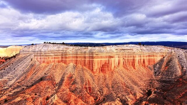 Wide view of eroded red sandstone mesa under dramatic cloudy sky, Spain. Flat topped layered plateau, orange and grey rock band, sculpted foreground badland, sparse shrub.