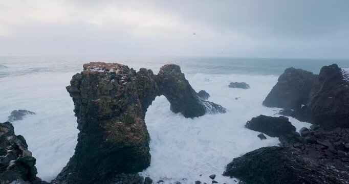 View of the Snaefellsnes stone arch with powerful waves crashing against dark volcanic rocks under an overcast sky in Eyja- og Miklaholtshr, Iceland.