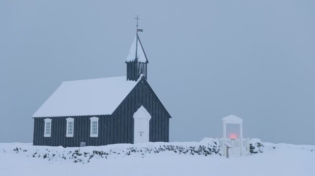 View of the historic black wooden church Budakirkja covered in thick snow with a stone wall under a grey winter sky in Eyja- og Miklaholtshr, Iceland.