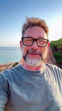 A happy man with glasses and a beard smiles at the camera, points, then continues to smile joyfully with a beautiful sea, cliff, and blue sky in the background, capturing a cheerful selfie moment duri