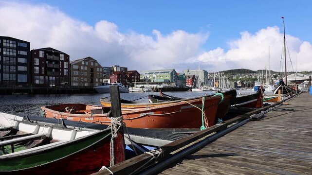 View of traditional wooden boats moored at a pier along the Nidelva riverfront with historic buildings under a blue sky in Trondheim, Trondelag, Norway.